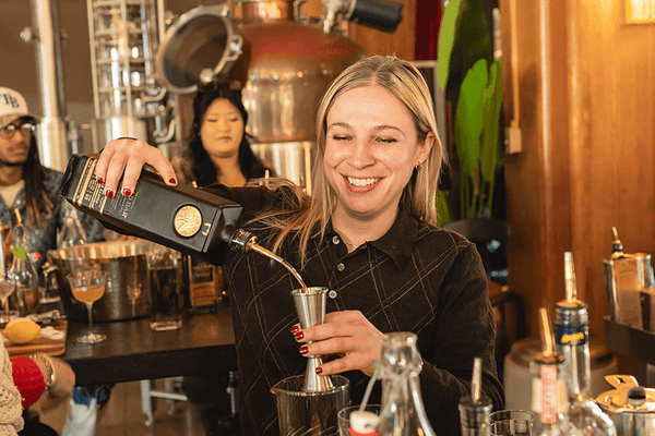 Woman in Gold Bar's Cocktail Class in San Francisco. She is pouring a drink, surrounded by bartending equipment and ingredients.